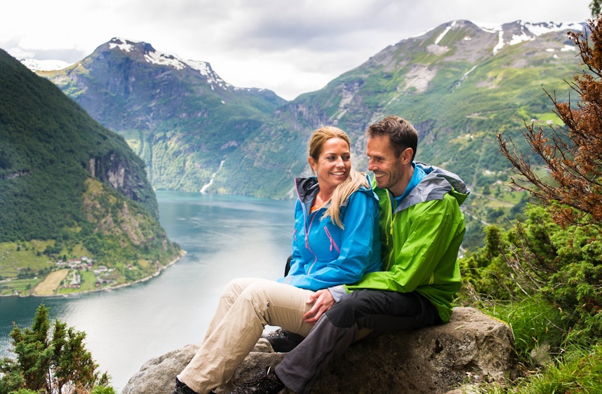 Geiranger Høst Ung mann og kvinne hviler seg på en stein. I bakgrunnen er det utsikt over Geirangerfjorden omkranset av fjell.